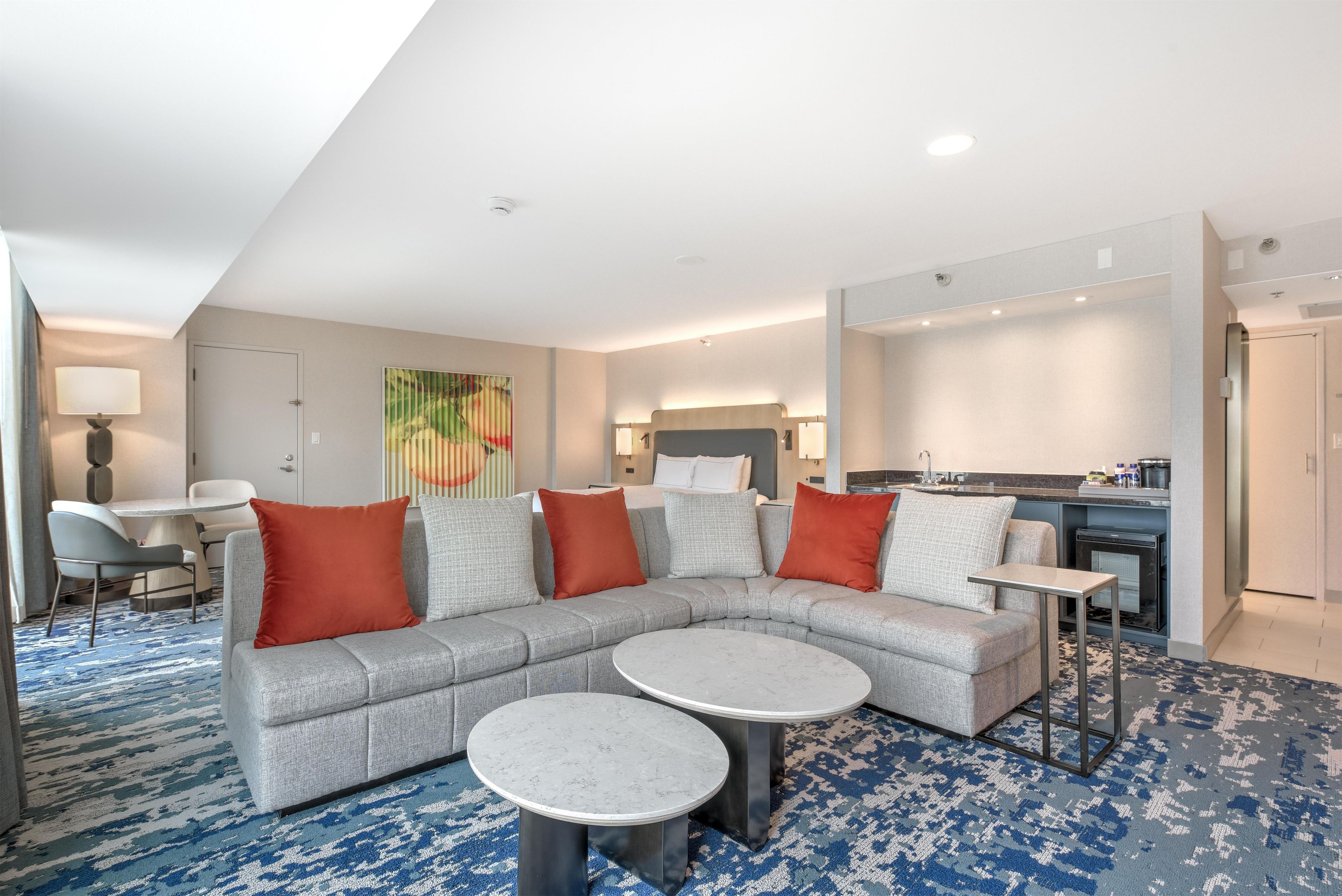 A modern hotel room with a sectional sofa, two round tables, a dining area, and a kitchenette.