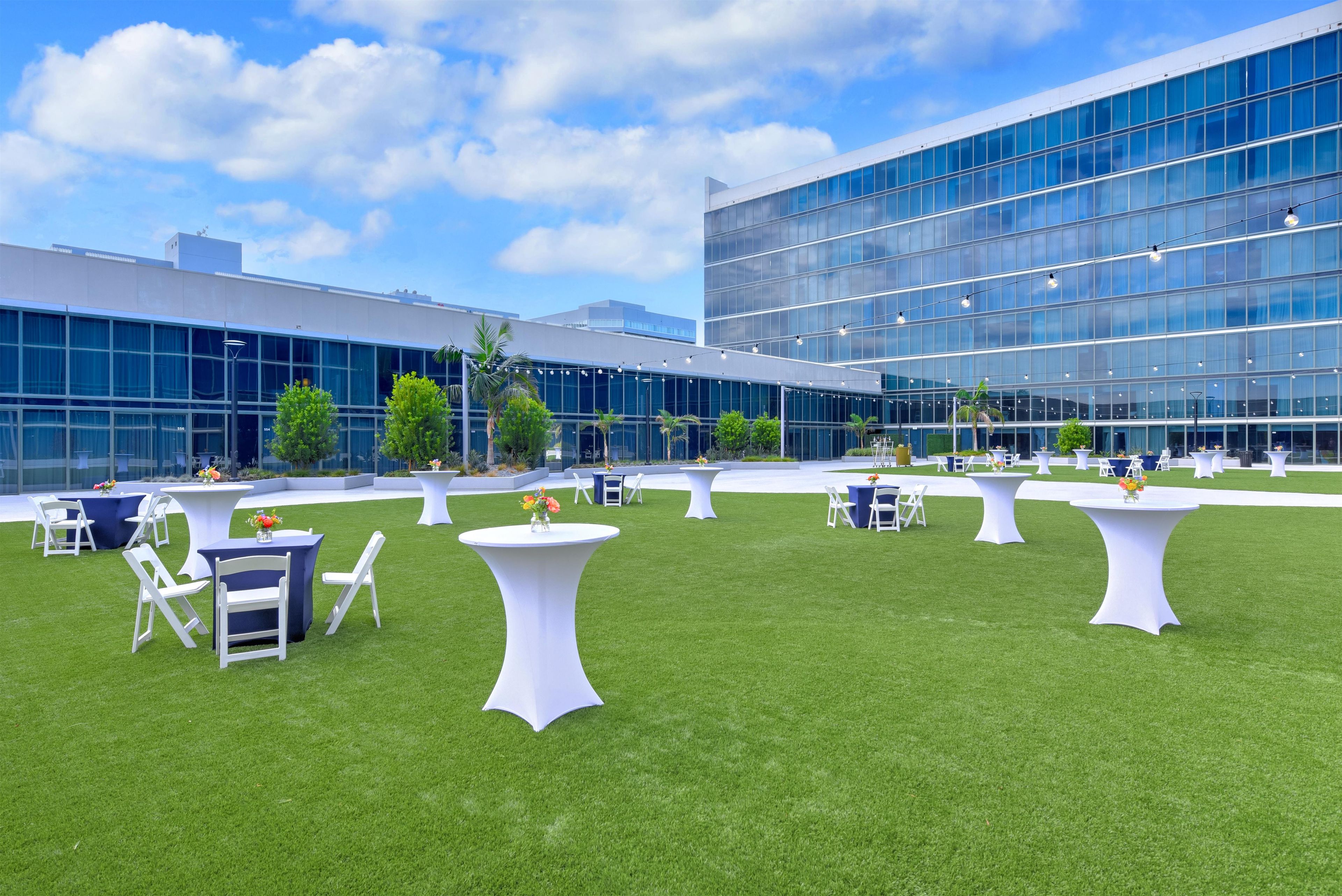 An outdoor event space with tables and chairs, a grassy lawn, and a modern building with large windows in the background.