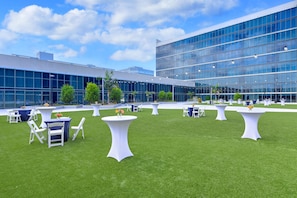 Un espacio para eventos al aire libre con mesas y sillas, un césped y un edificio moderno con grandes ventanales al fondo.