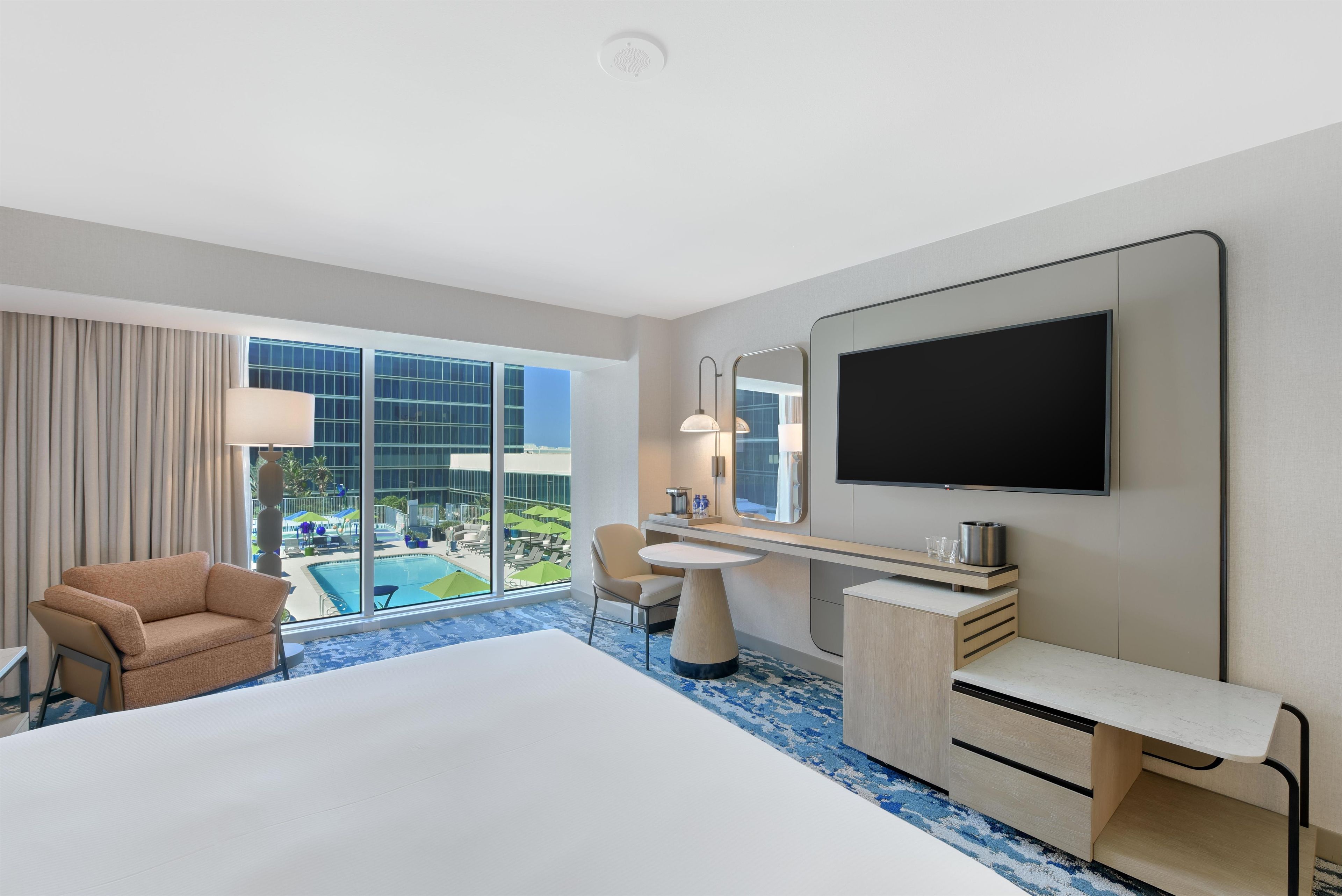 A hotel room with a large bed, a desk, a chair, a TV, and a view of a pool and building.
