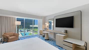 A hotel room with a large bed, a desk, a chair, a TV, and a view of a pool and building.