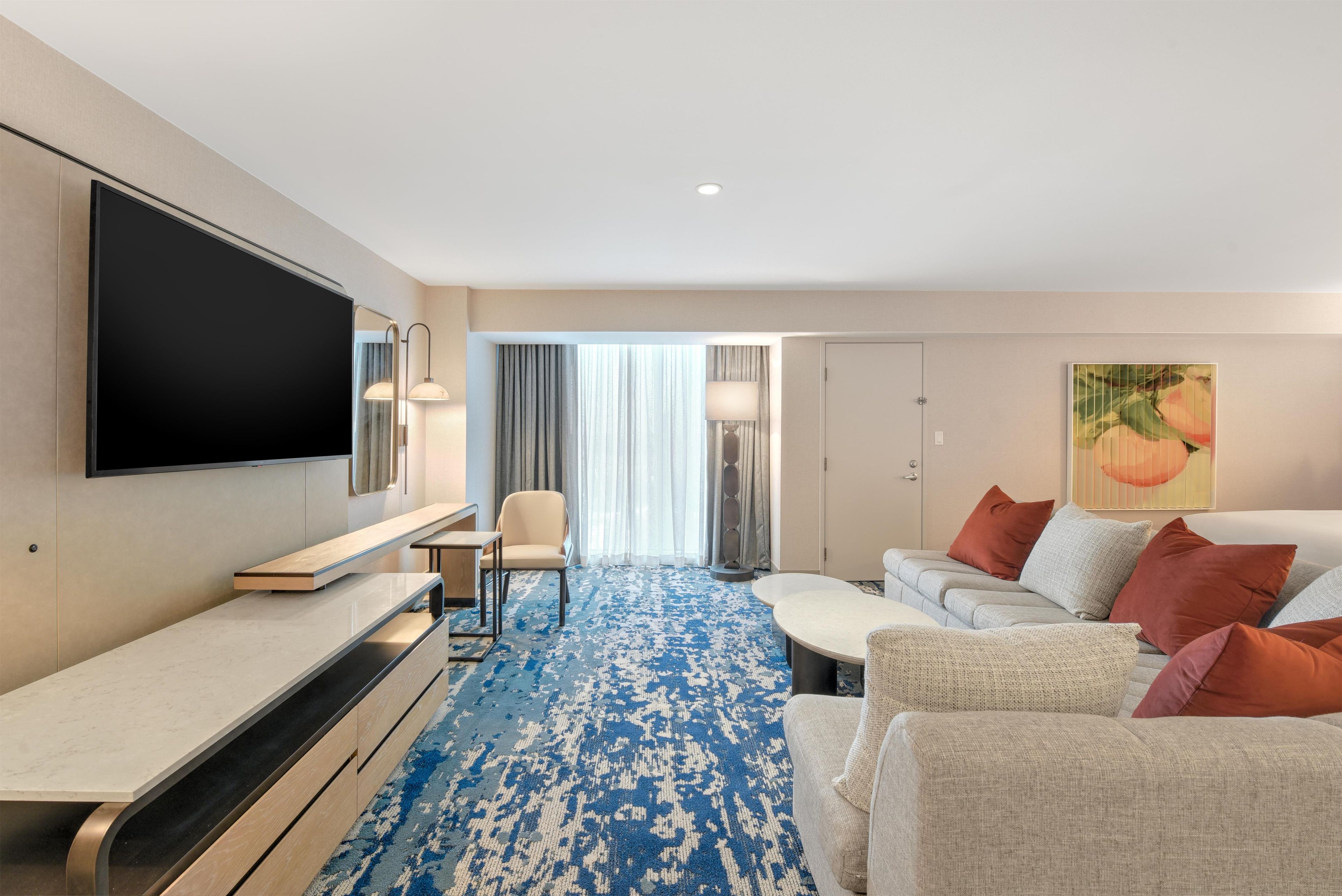 A hotel room with a flat-screen TV, a sofa, a coffee table, a chair, and a patterned carpet.