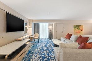 A hotel room with a flat-screen TV, a sofa, a coffee table, a chair, and a patterned carpet.