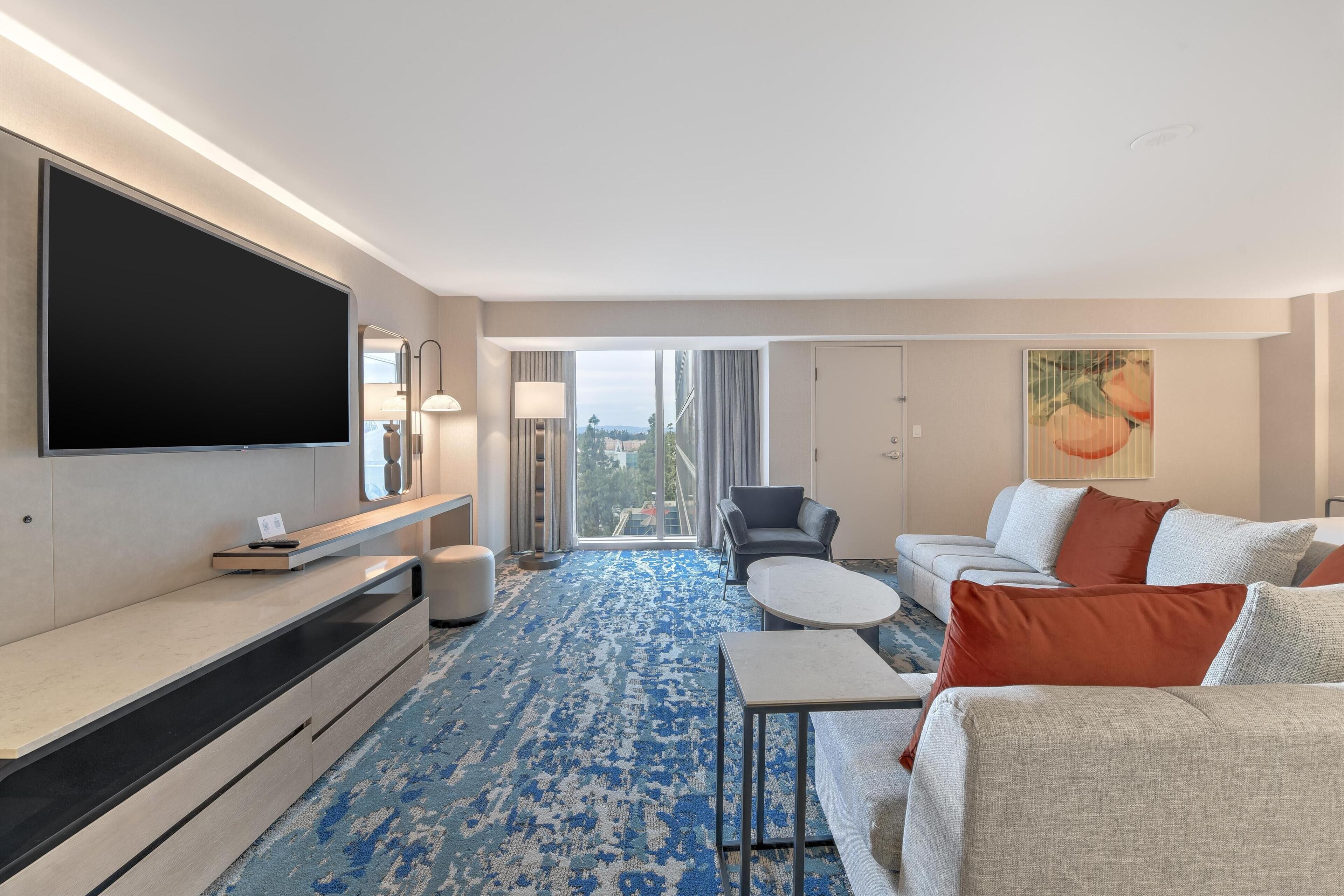 A modern hotel room with a large flat-screen TV, a sofa set, a coffee table, and a patterned carpet.