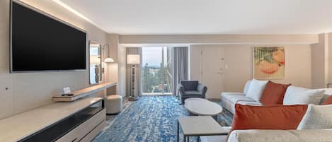 A modern hotel room with a large flat-screen TV, a sofa set, a coffee table, and a patterned carpet.