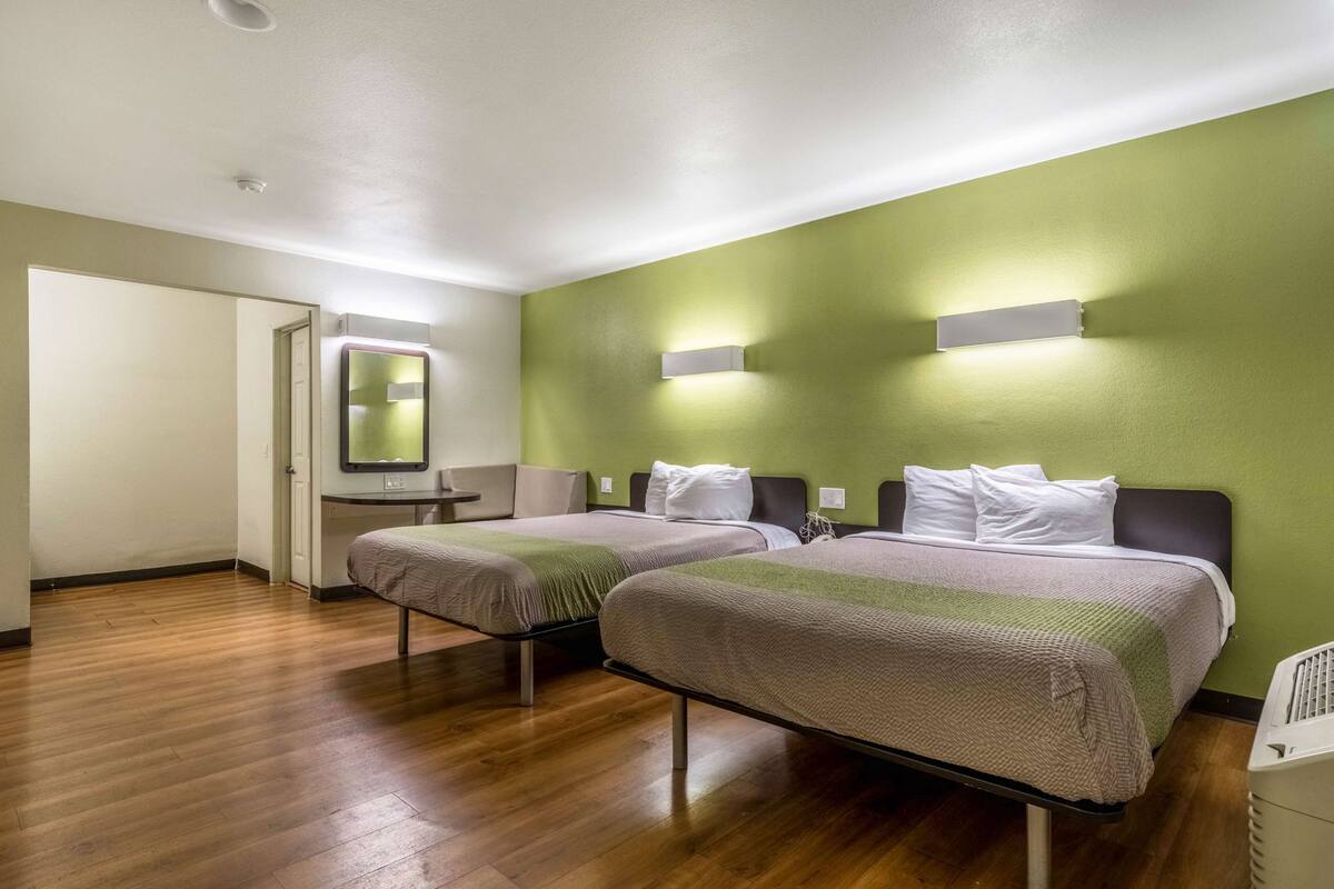 A hotel room with two beds, a wooden floor, a green wall, and a mirror above the door.