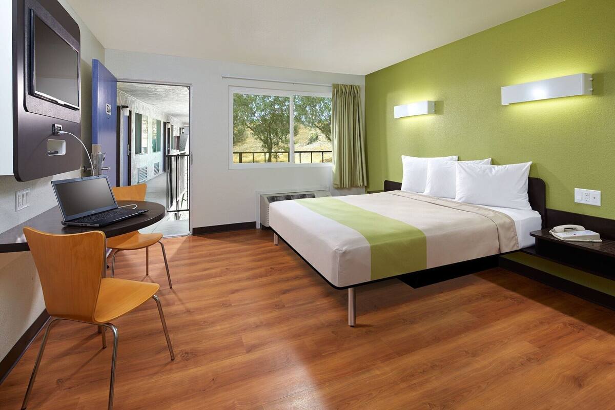 A hotel room with a bed, a desk with a laptop, a chair, and a window with a view of the outdoors.