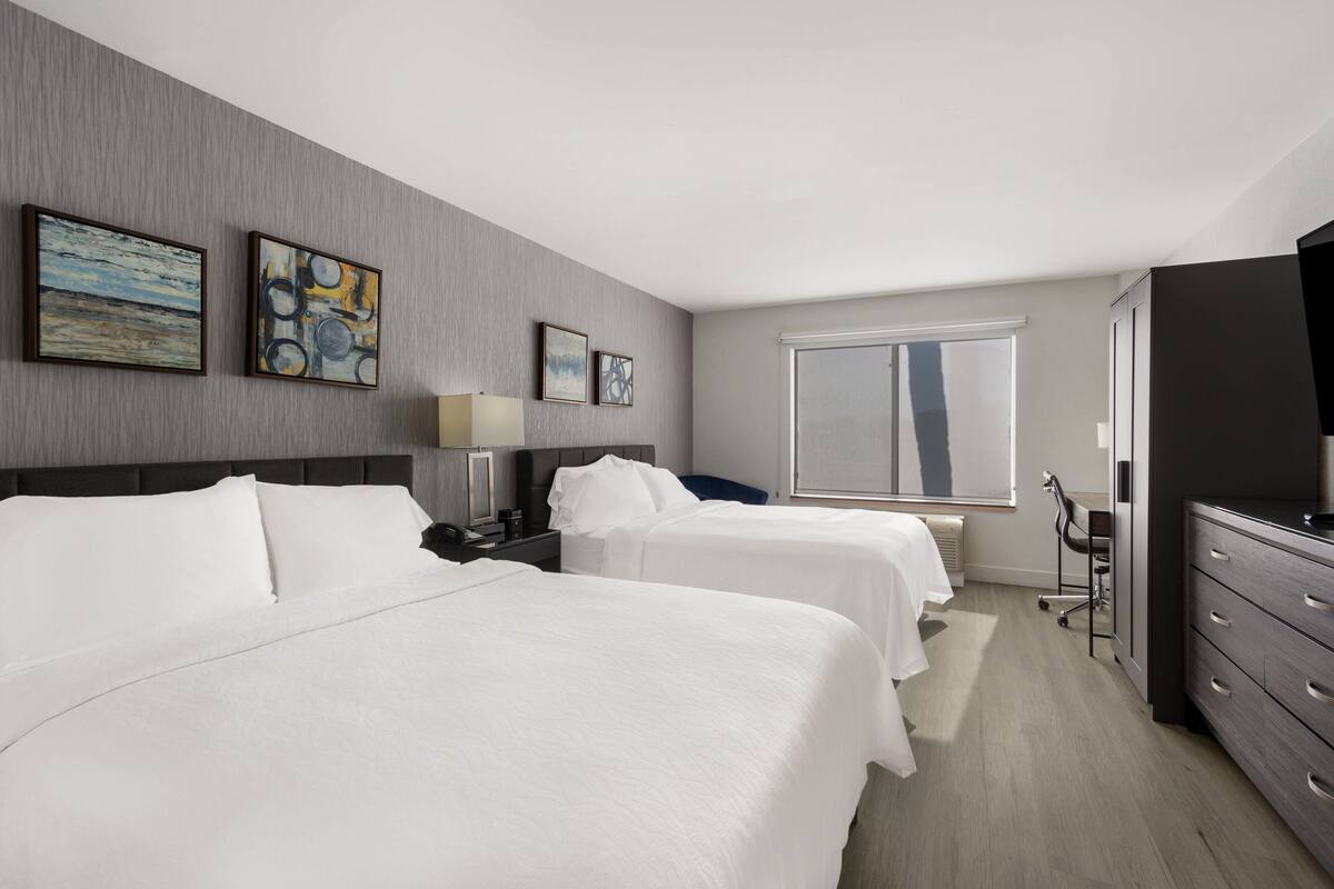 A hotel room with two beds, a desk, a chair, a TV, and artwork on the wall.