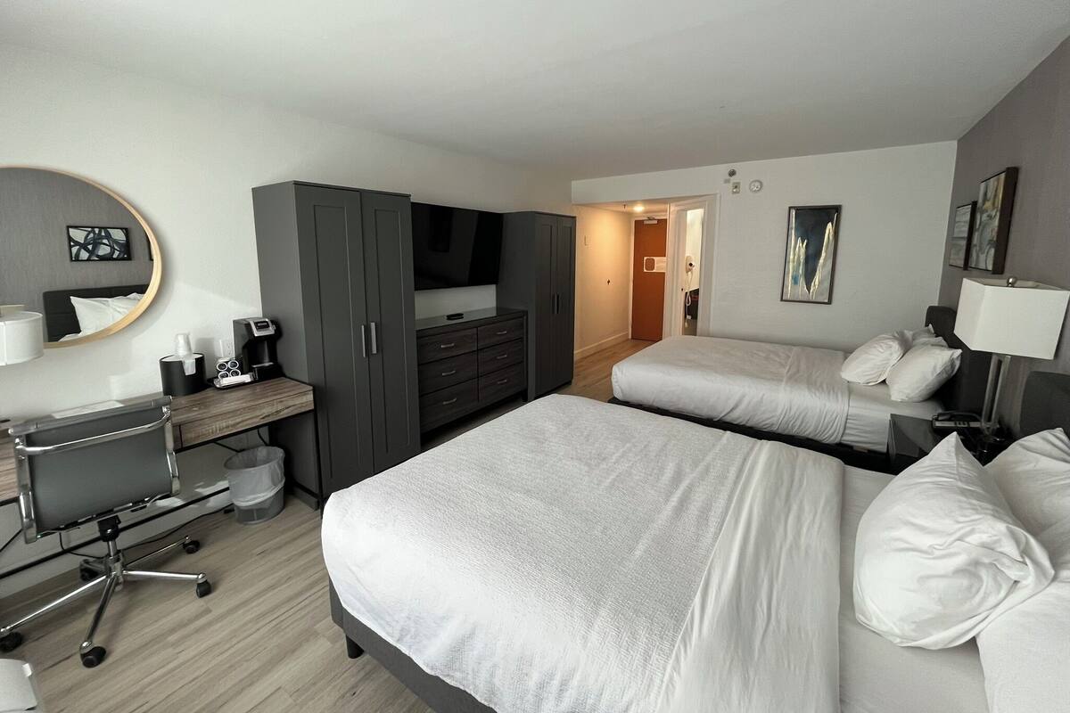 A hotel room with two beds, a desk with a chair, a wardrobe, a TV, and a bathroom visible through an open door.