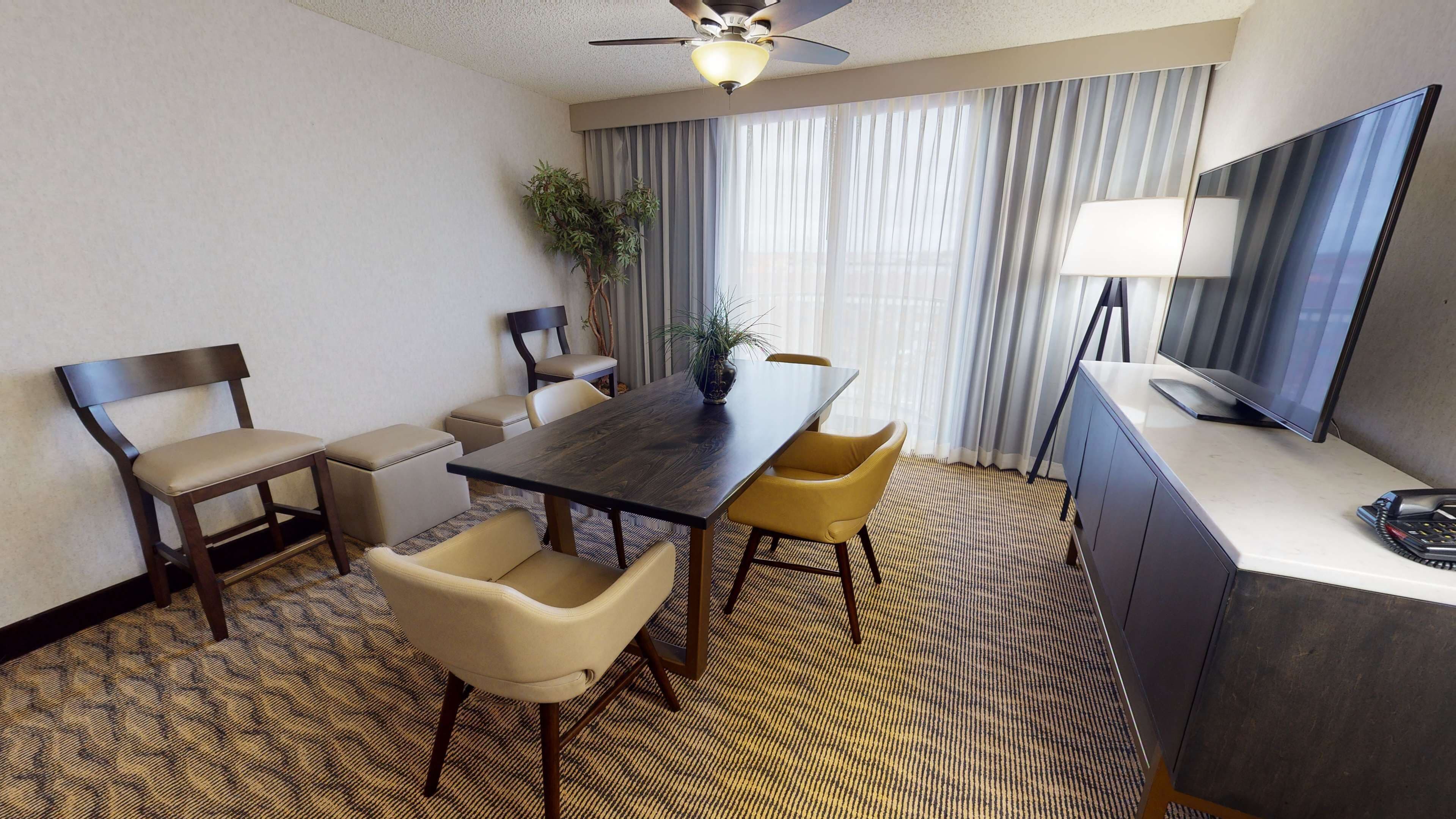 Habitación de hotel con una mesa de comedor de madera oscura, sillas color beige y amarillo, una pequeña planta en maceta, un televisor y un escritorio con una lámpara.