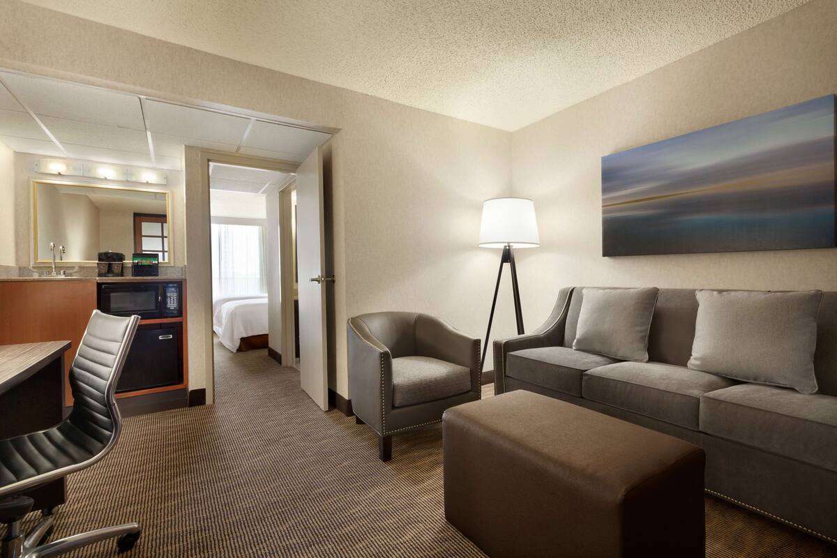 Ein Hotelzimmer mit Schreibtisch, Stuhl, Sofa, Hocker, Lampe, einem Gemälde und einem Bett im Hintergrund.