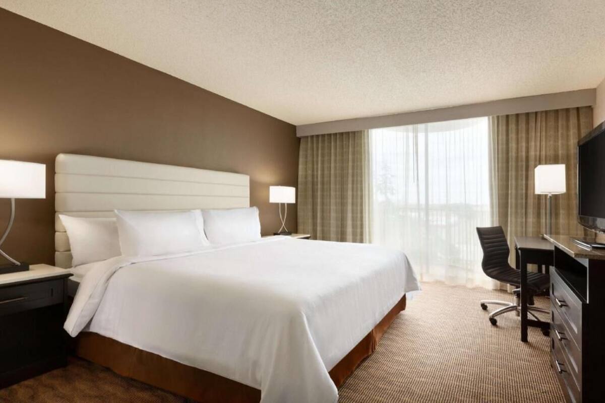 Ein Hotelzimmer mit einem großen Bett, einem Schreibtisch mit Stuhl, einem Fernseher und einem Fenster mit Vorhängen.