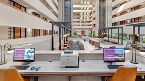Un amplio vestíbulo de hotel con varias zonas de estar, una escalera central y una lámpara de araña moderna. Hay dos puestos de trabajo con computadoras e impresoras.