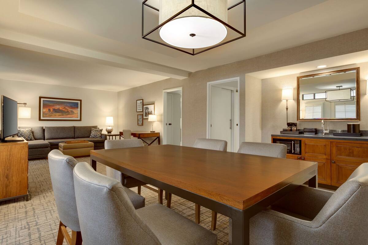A spacious living room with a large wooden dining table, grey upholstered chairs, a grey sofa, and a television. There is a kitchen area with a microwave, coffee maker, and a large mirror above the counter.