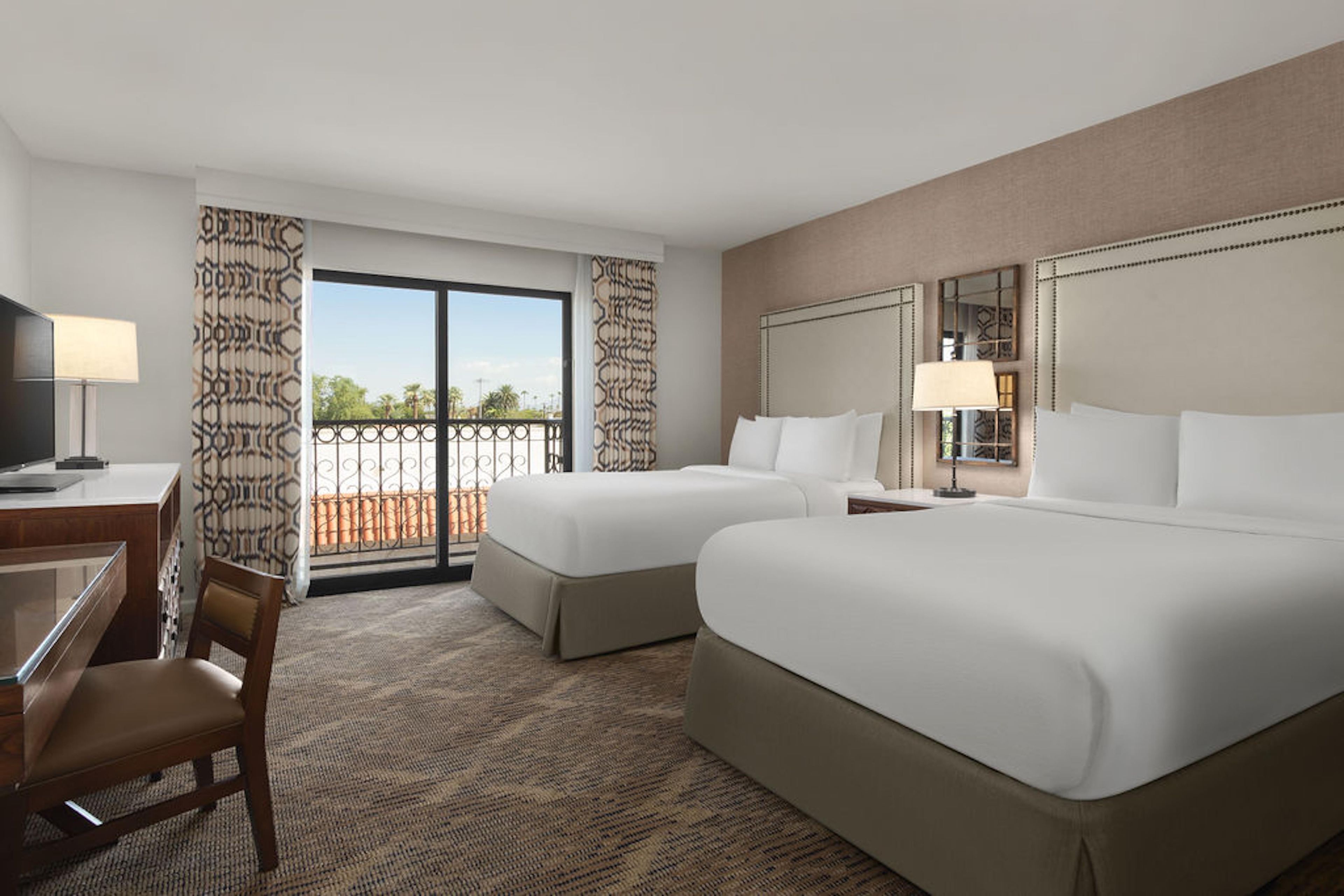 Quarto de hotel com duas camas, uma escrivaninha com cadeira, televisão e varanda com cortinas estampadas.