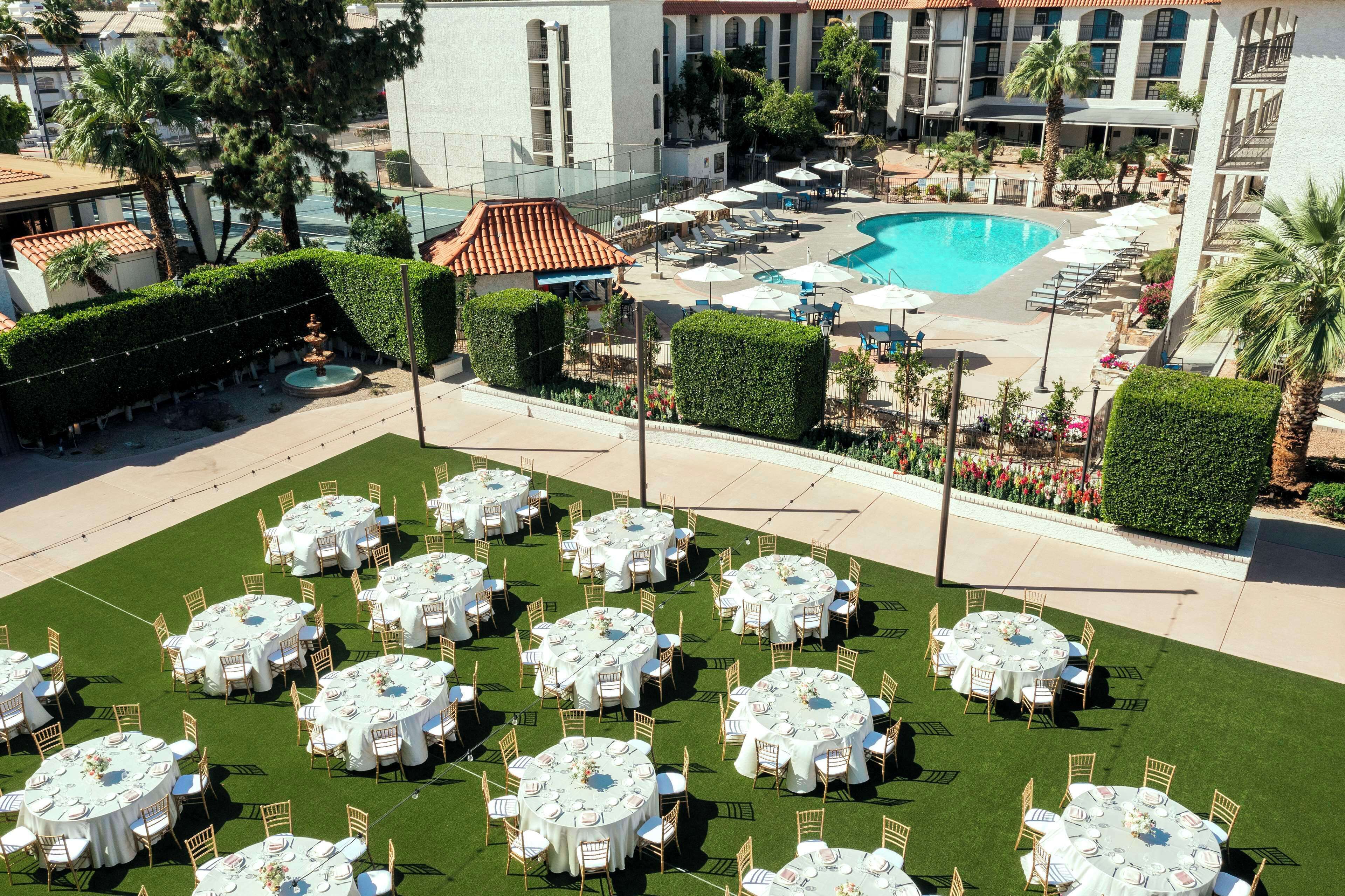 Un pati d'hotel amb diverses taules rodones preparades per dinar, una piscina i una zona de jardí paisatjística.