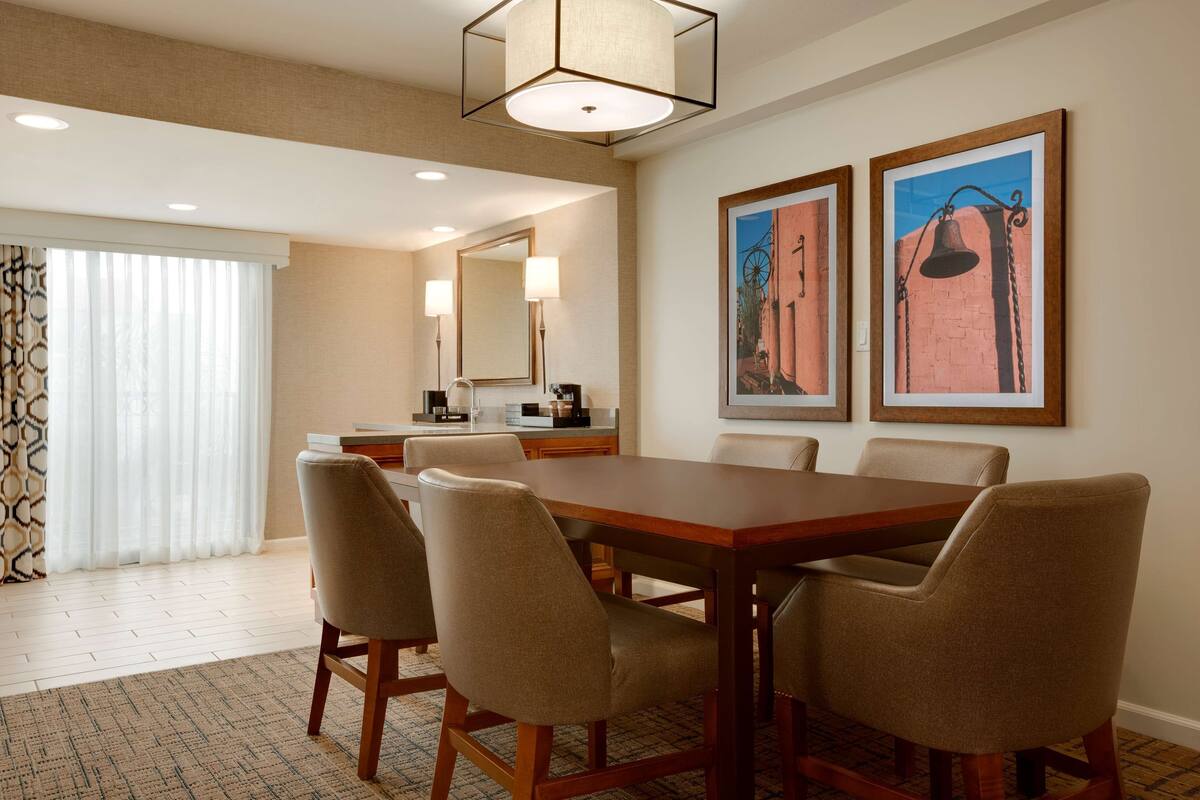A hotel dining area with a large wooden table, four beige chairs, a chandelier, and two framed pictures on the wall.