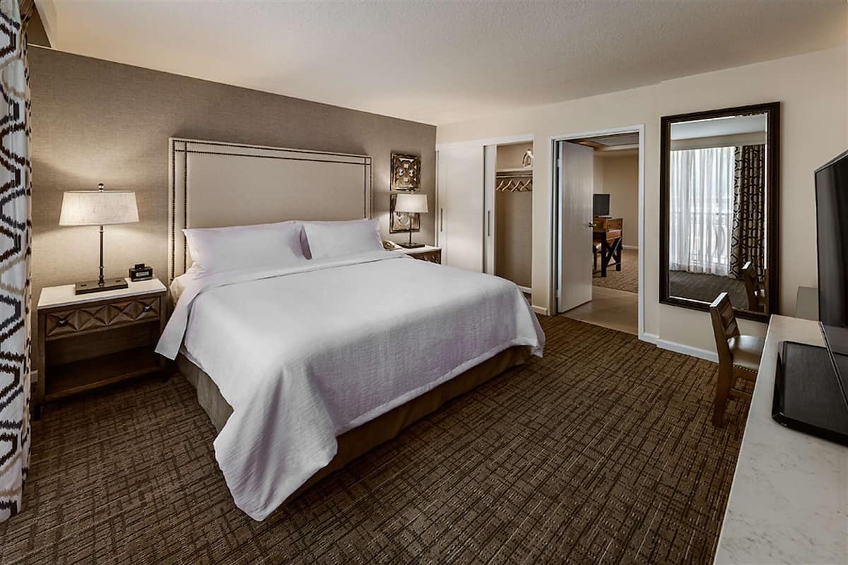 A hotel room with a large bed, bedside tables with lamps, a desk with a chair, a wardrobe, and a mirror.