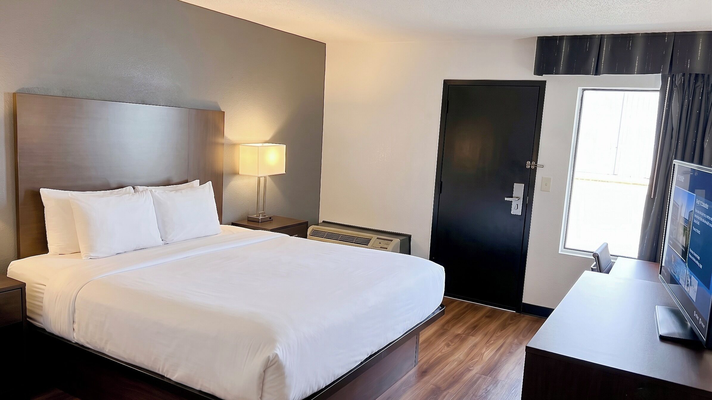 Una habitación de hotel con una cama grande, una lámpara de noche, una puerta y un escritorio con una computadora.