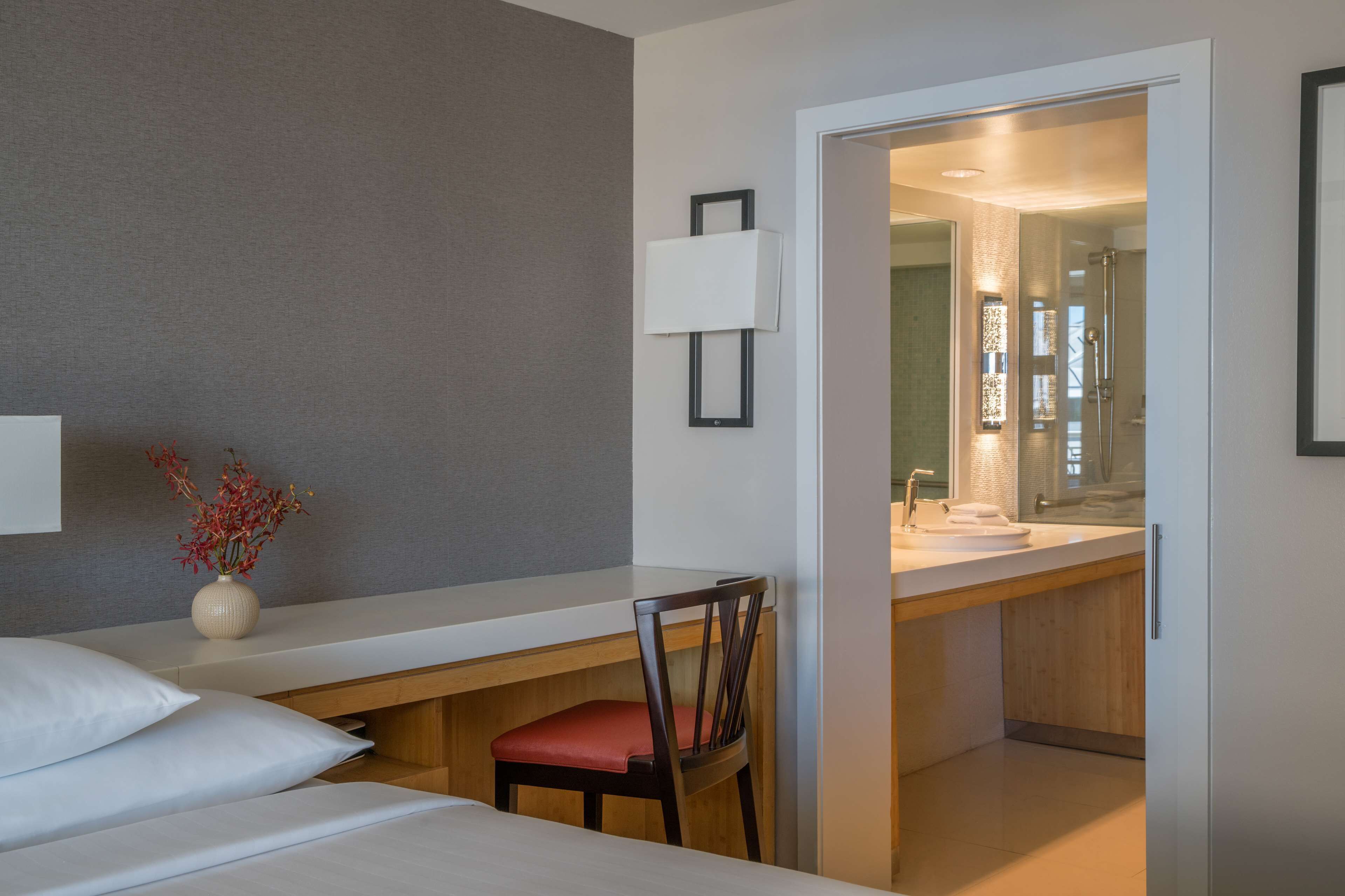 A hotel room with a bed, a desk, a chair, a vase with flowers, a wall-mounted lamp, and a bathroom with a sink and mirror.