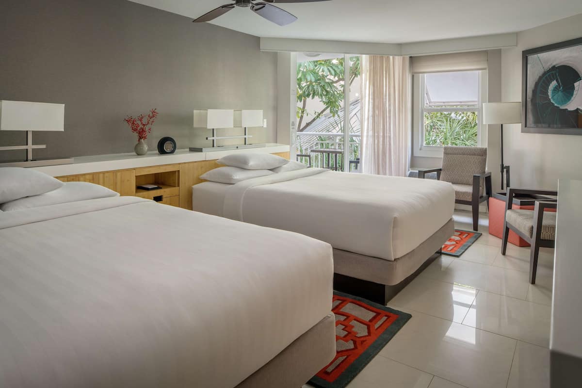 A hotel room with two beds, a desk with a chair, a window with a view of greenery, and a ceiling fan.