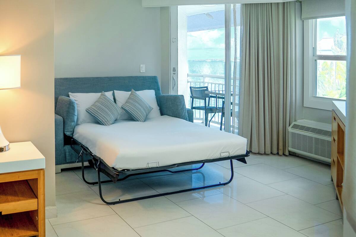 A single bed with a metal frame, a bedside table with a lamp, and a view of the outdoors through a large window.