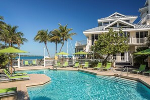 A pool area with lounge chairs and umbrellas, surrounded by palm trees and a building with balconies.