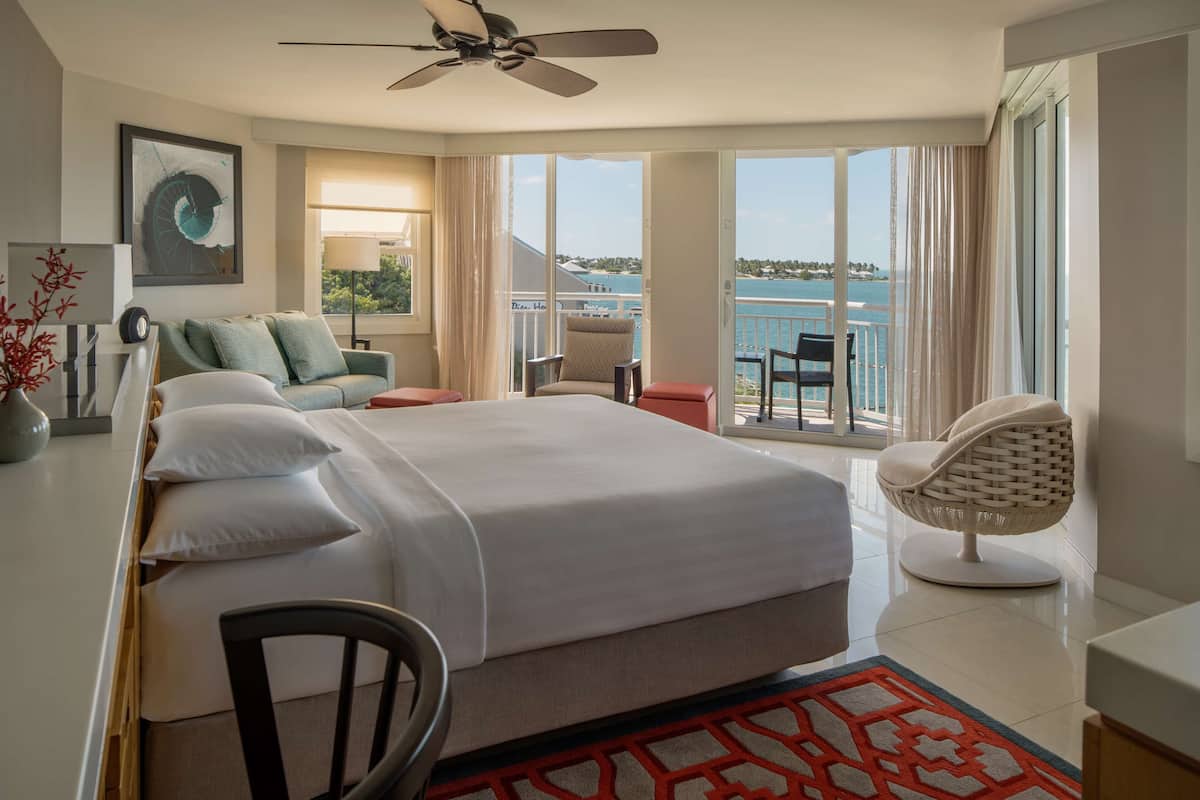 A hotel room with a large bed, a ceiling fan, a view of the sea, a balcony with a chair, and a desk with a lamp.