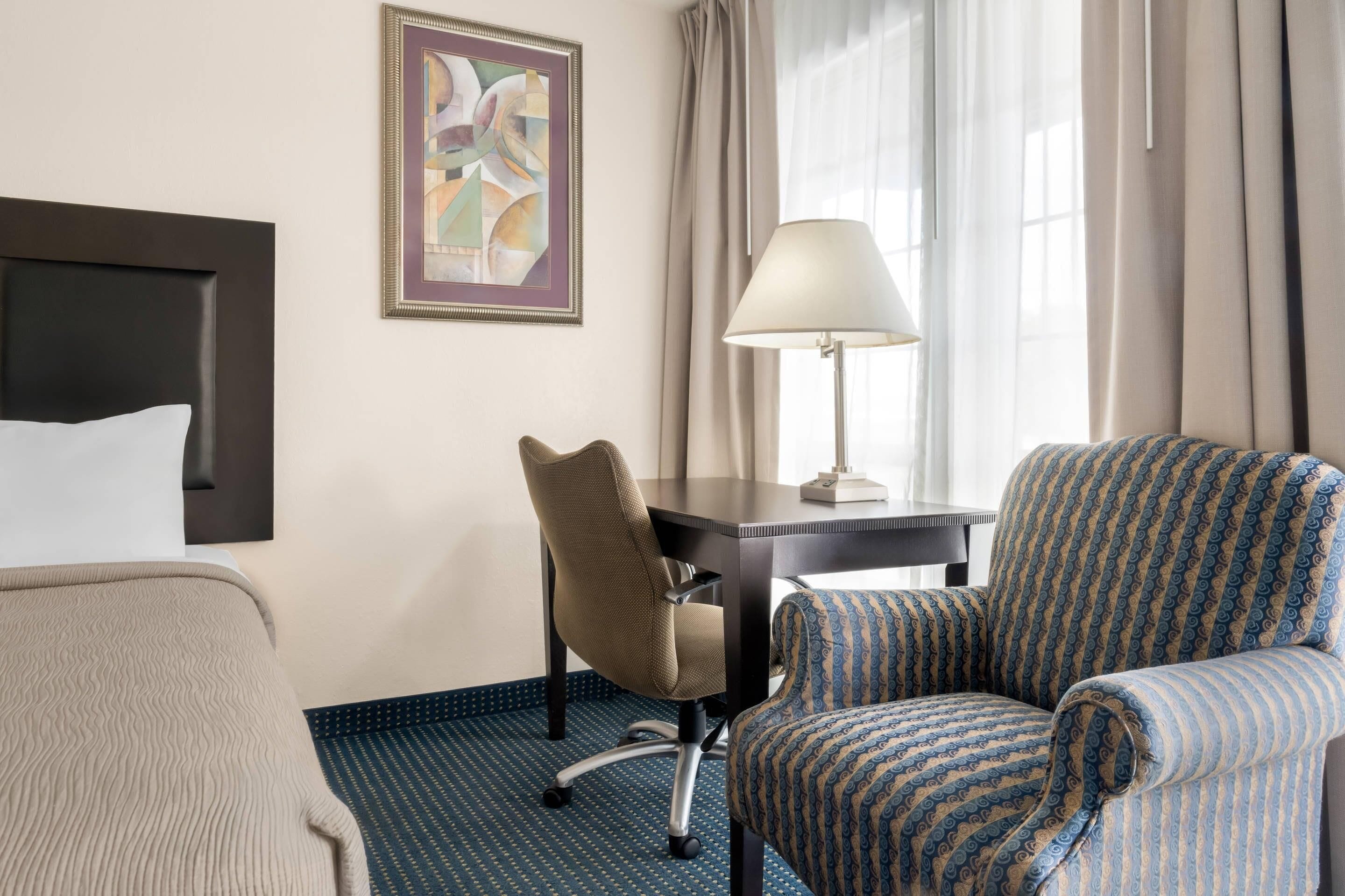 A hotel room with a bed, a desk, a chair, a lamp, and a painting on the wall.