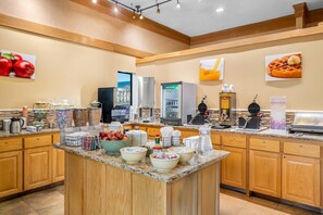 A hotel breakfast area with a central island, various food items, and a refrigerator.