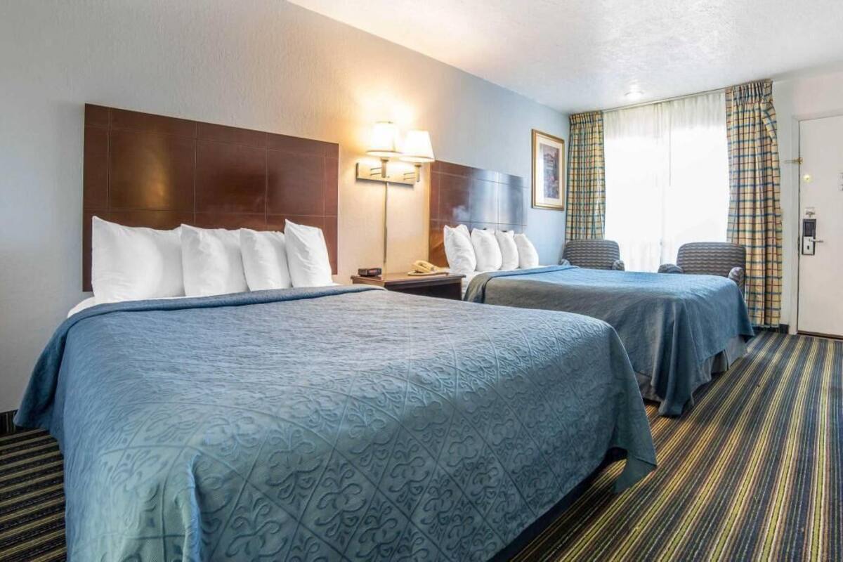 A hotel room with two beds, a nightstand with a lamp, a chair, and a door with curtains.