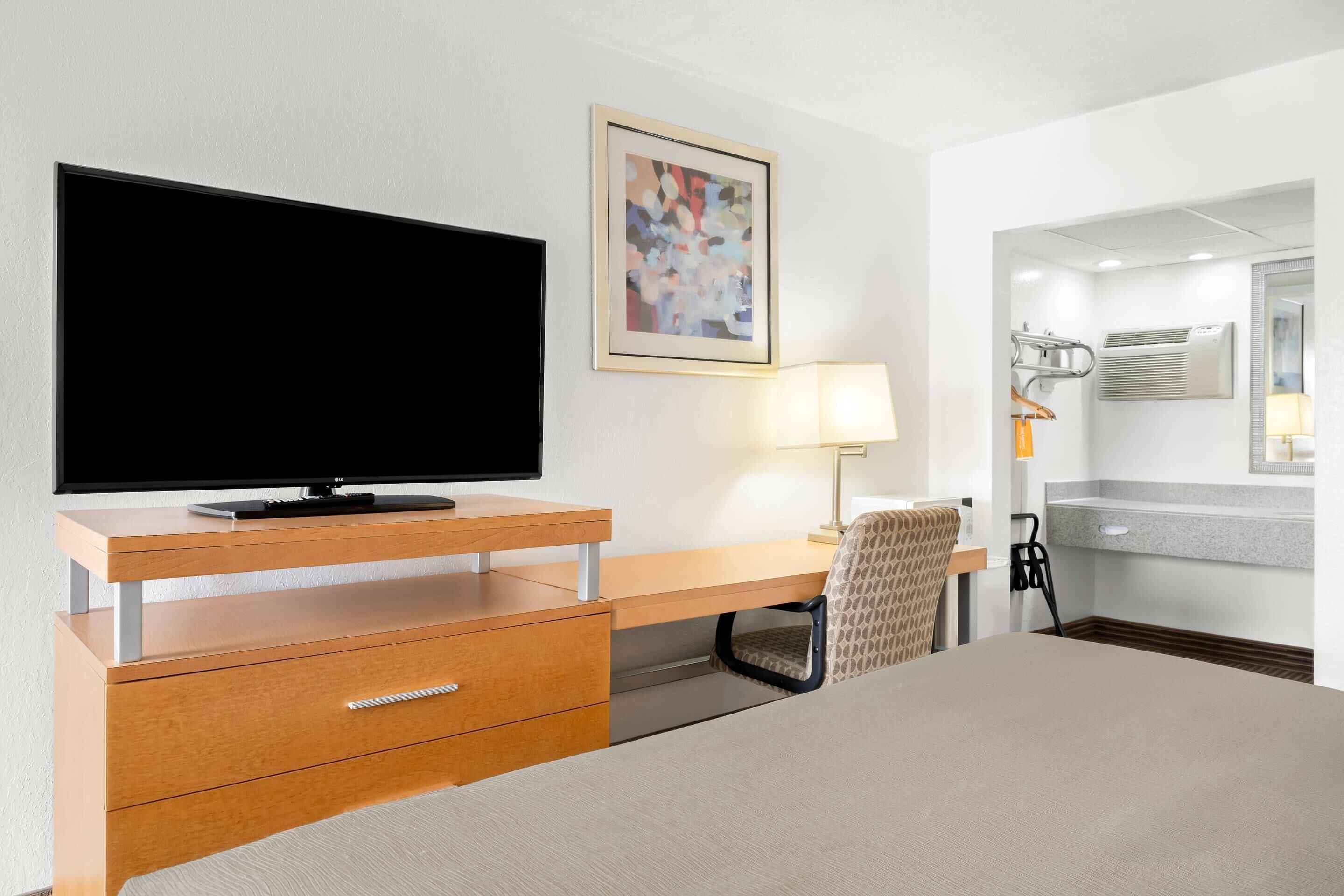 A hotel room with a flat-screen TV, a desk with drawers, a chair, a bedside table, a lamp, a mirror, and a bathroom with a shower and toilet.