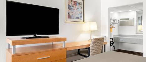 A hotel room with a flat-screen TV, a desk with drawers, a chair, a bedside table, a lamp, a mirror, and a bathroom with a shower and toilet.