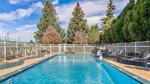 A rectangular swimming pool with clear blue water, surrounded by a white fence, and trees in the background.