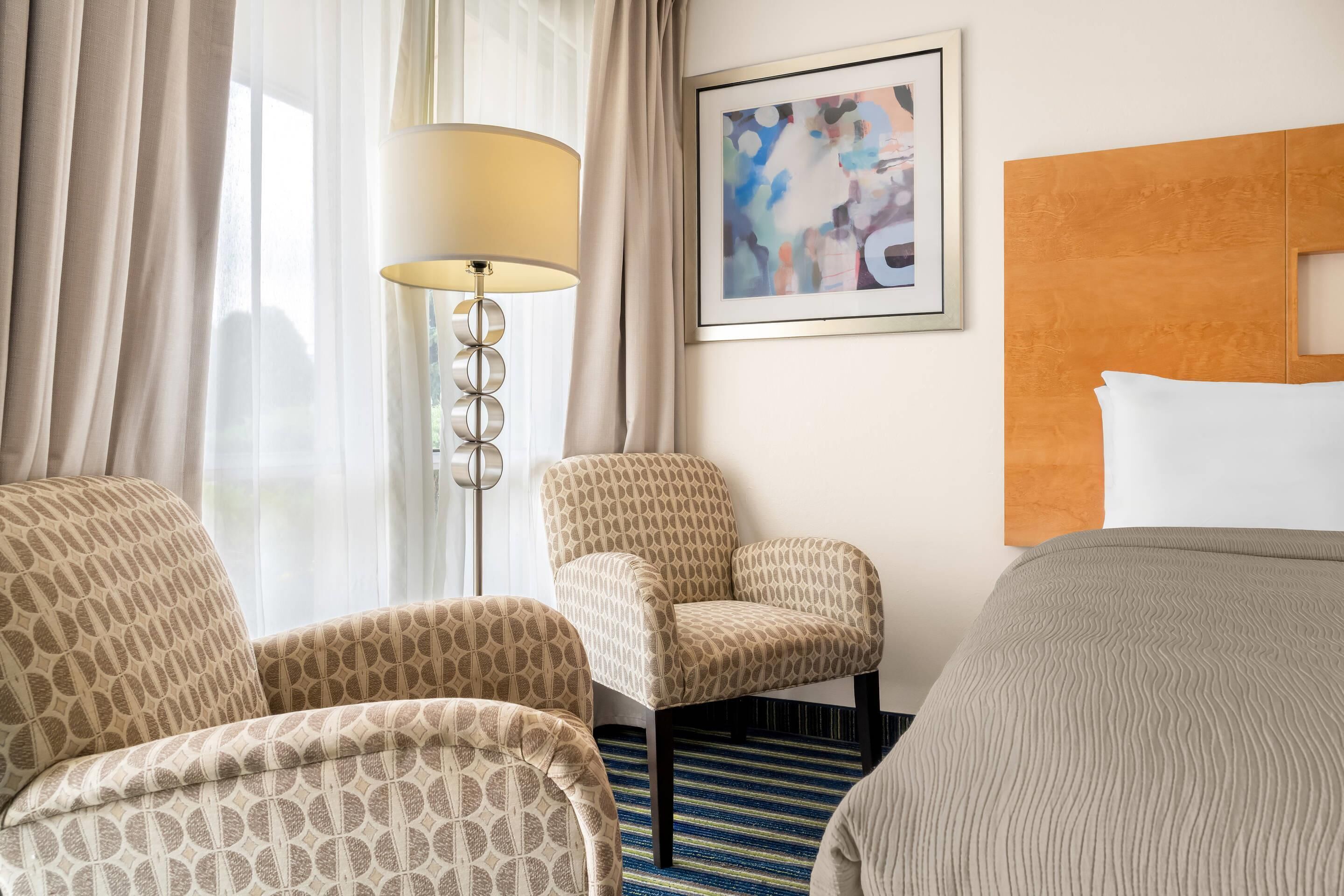 Une chambre d’hôtel avec un lit, une chaise, une lampe sur pied et un tableau au mur.