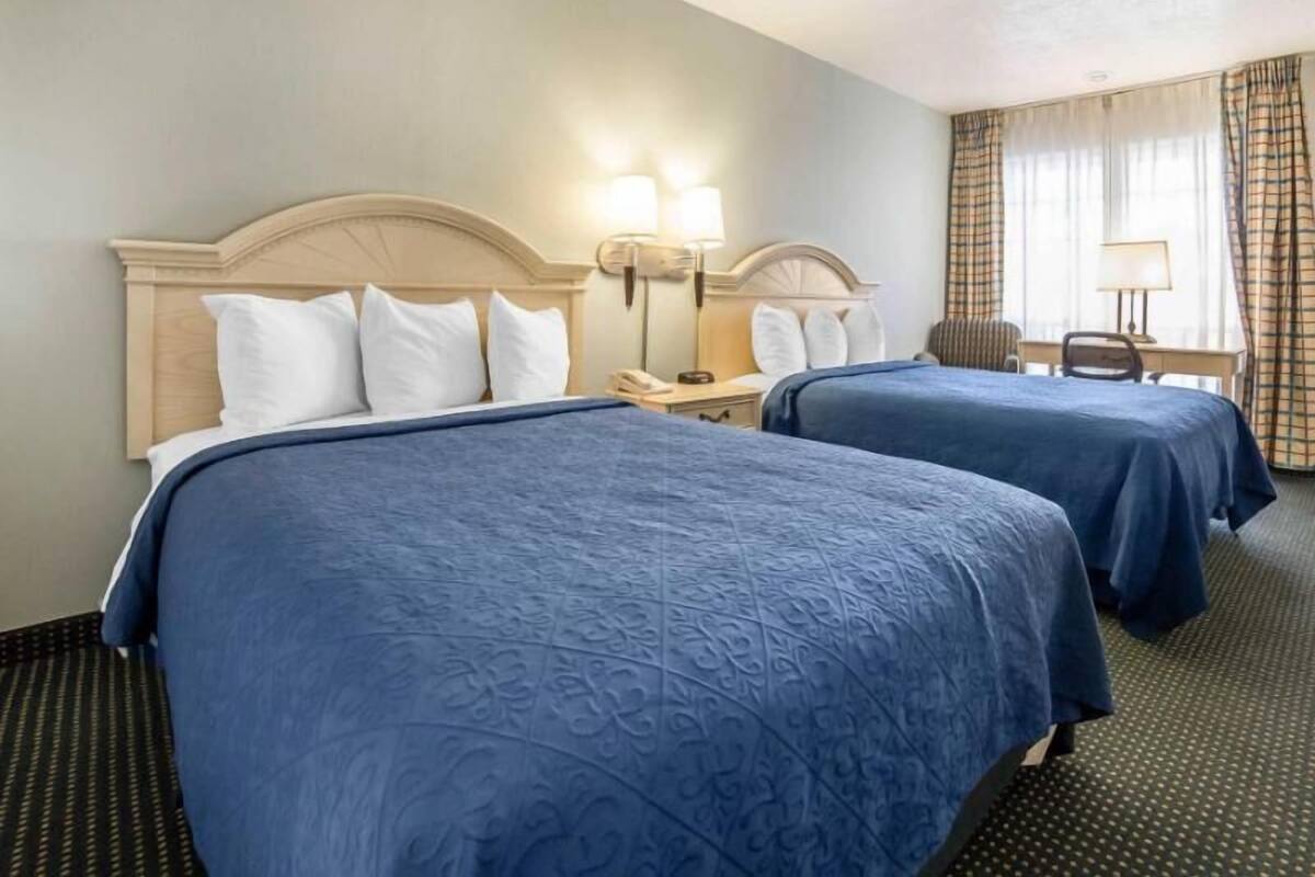 A hotel room with two beds, a desk, a chair, and a lamp.