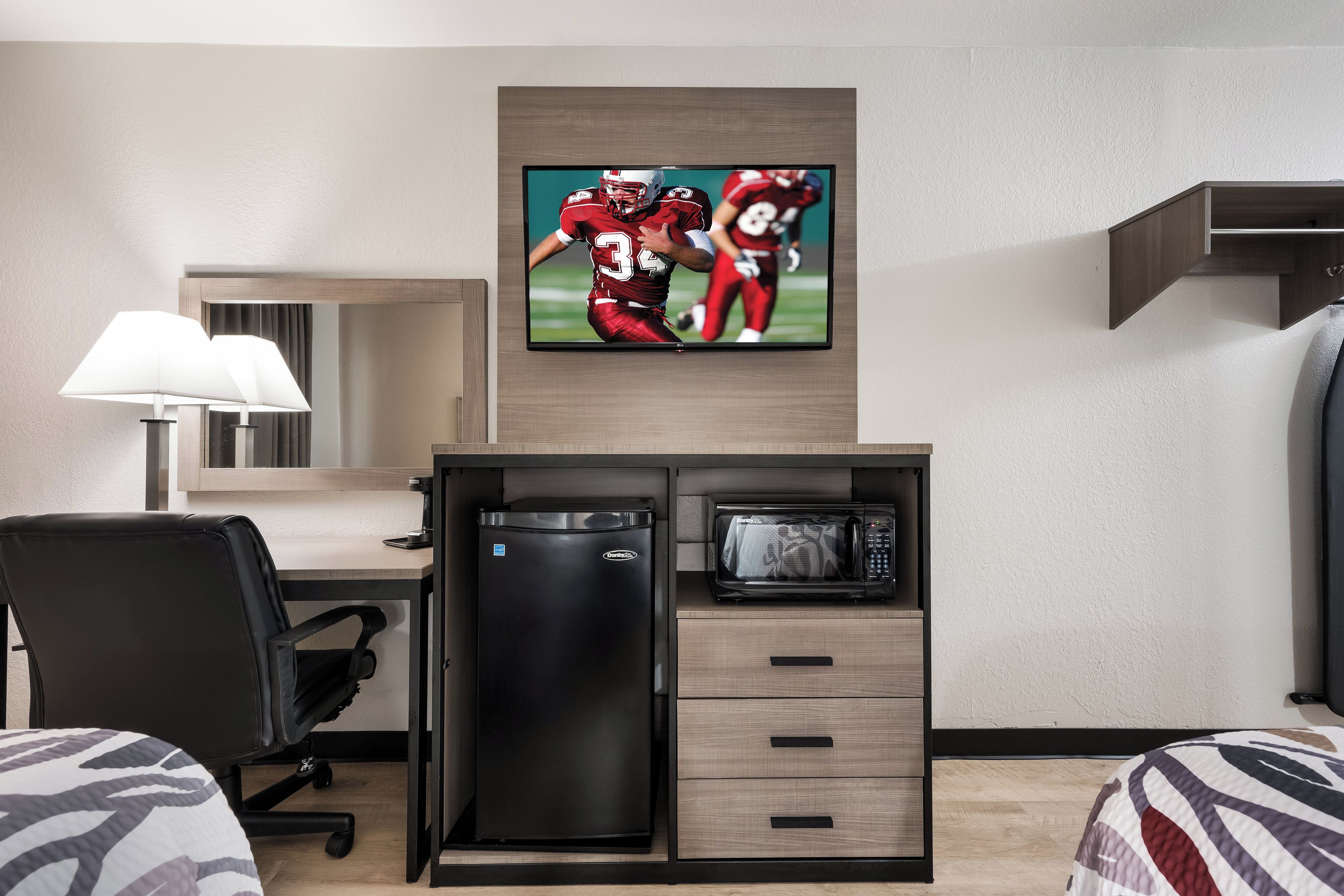 A hotel room with a flat-screen TV mounted above a desk, a microwave, a refrigerator, a chair, and a bed with striped bedding.