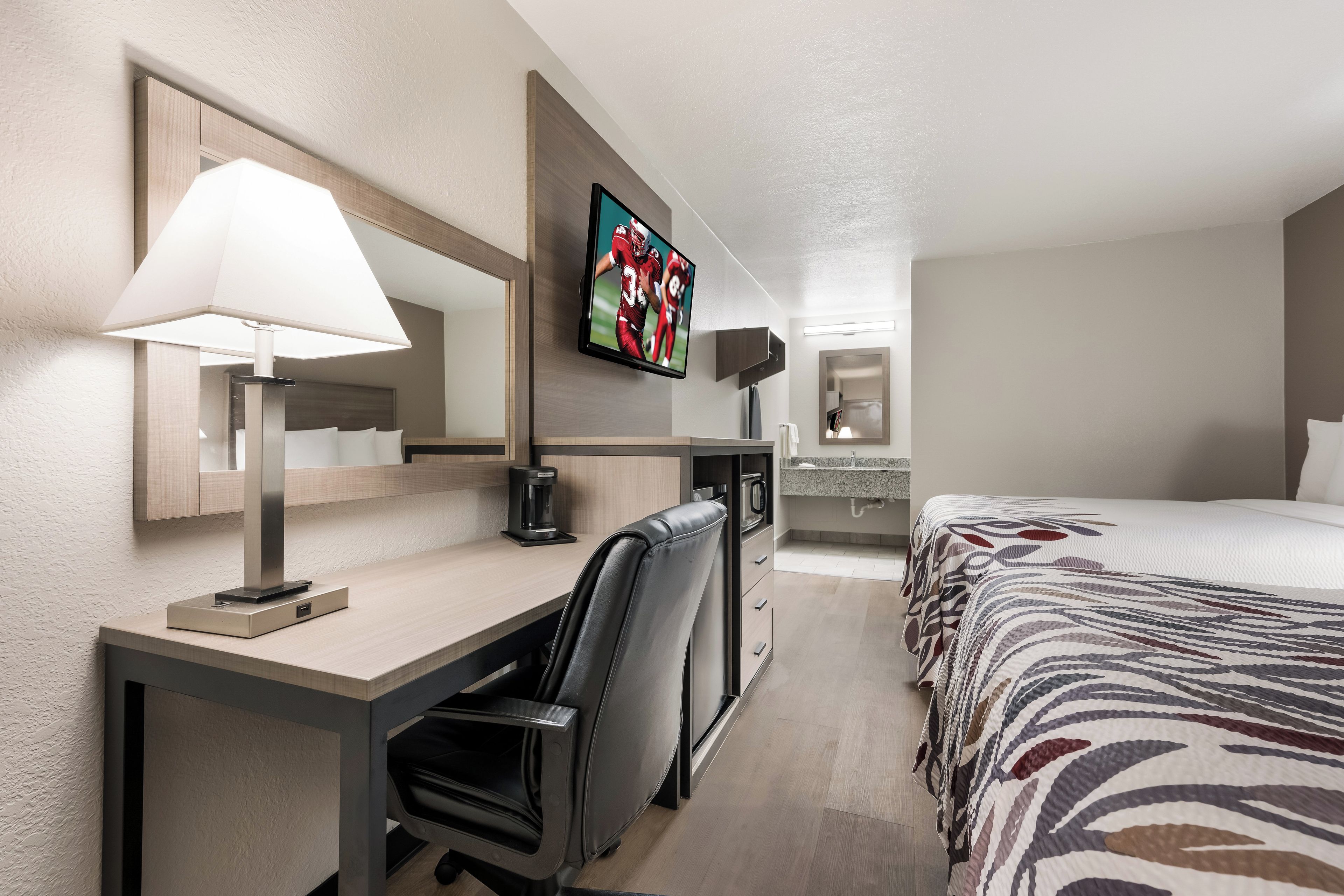 A hotel room with a desk, chair, bed, TV, and a coffee maker.