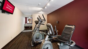 Un gimnasio con equipo de ejercicio, un televisor mostrando 'Wi-Fi gratuito en todas las habitaciones' y un espejo al fondo.
