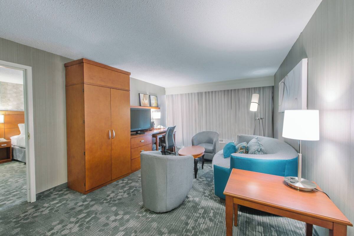 A hotel room with a wooden wardrobe, a grey armchair, a blue sofa, a small round table, and a lamp.