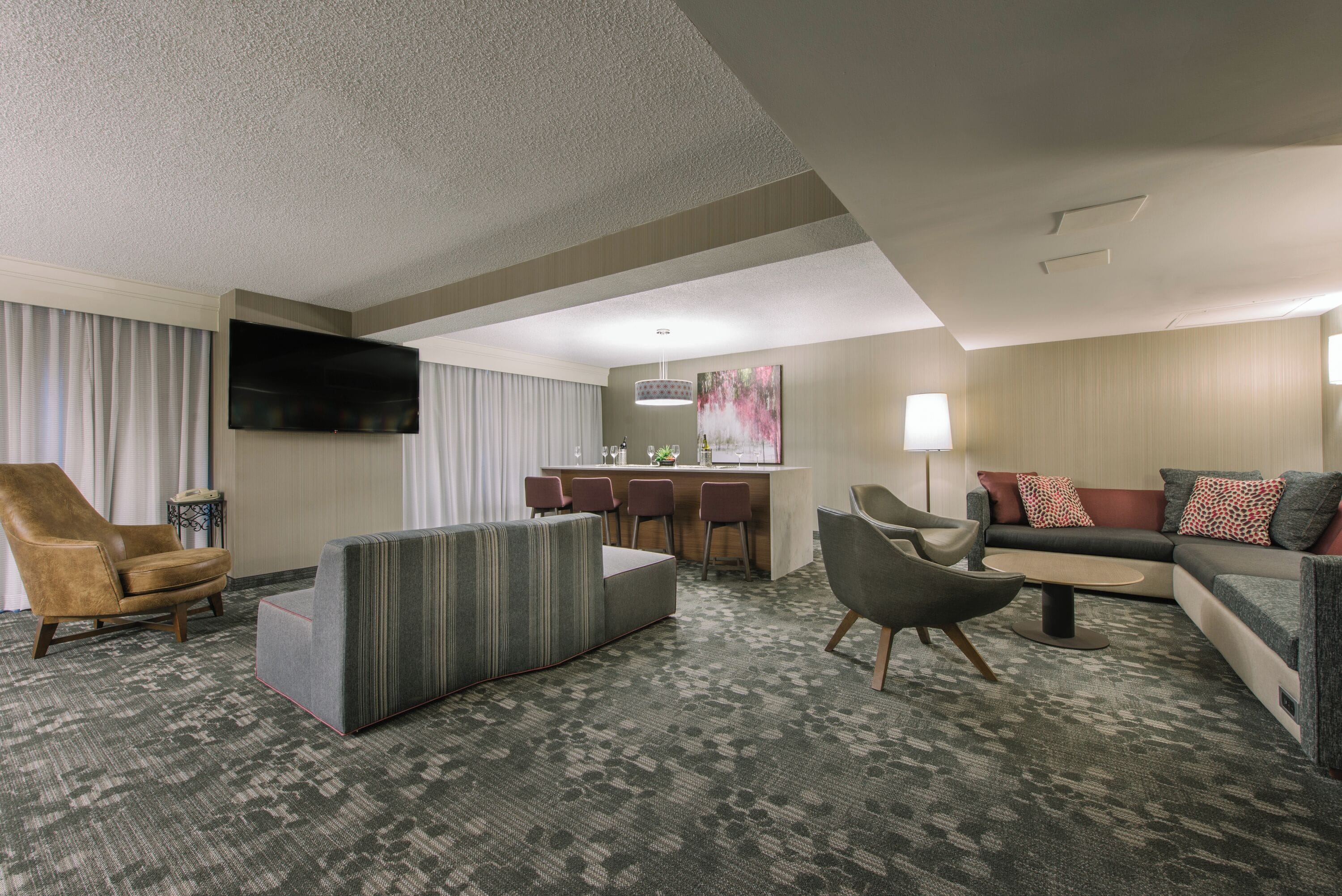 A hotel room with a sofa, armchair, ottoman, a flat-screen TV mounted on the wall, a small bar area with stools, and a lamp.