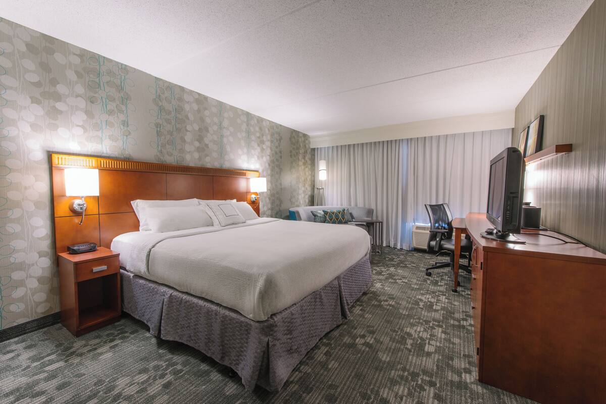 A hotel room with a large bed, a desk with a computer, a chair, a TV, and a nightstand with a lamp.