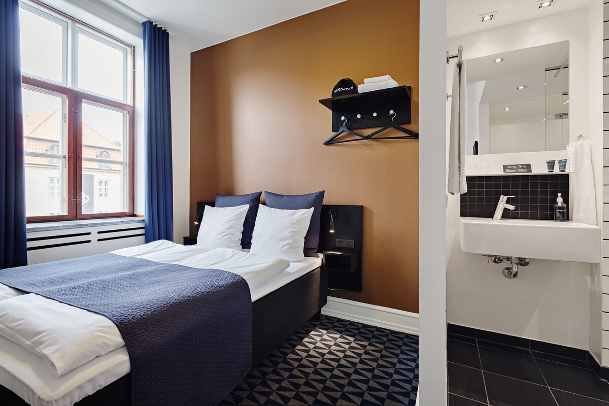 Une chambre d’hôtel comprenant un lit, une table de chevet, une étagère fixée au mur et une salle de bain avec un lavabo et un miroir.