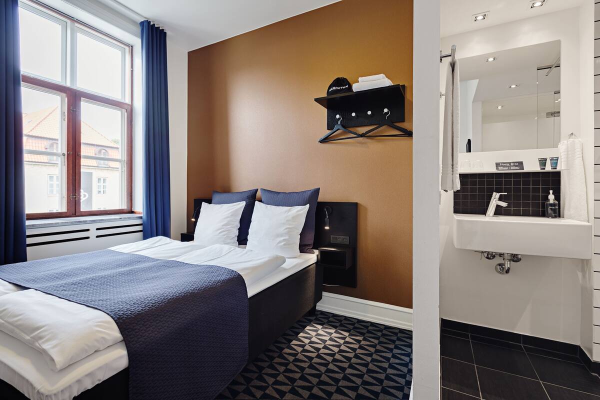 Een hotelkamer met een bed, nachtkastje, een wandplank en een badkamer met een wastafel en spiegel.