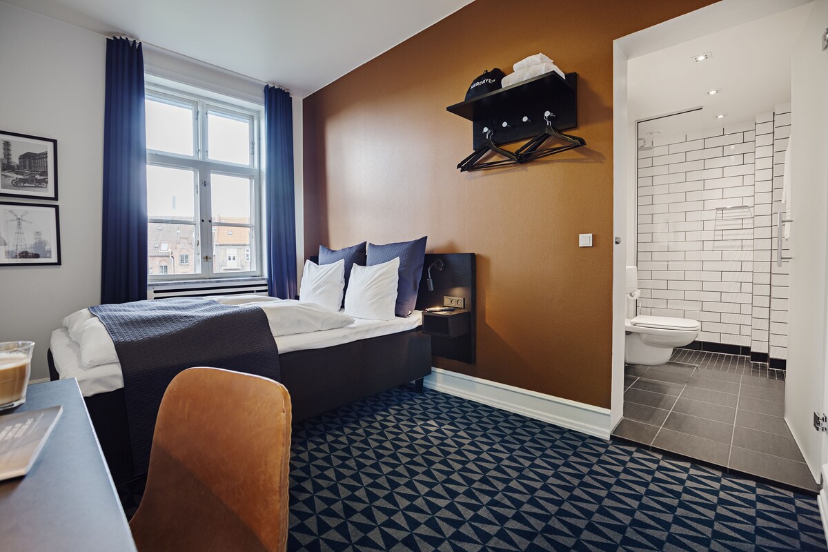 A hotel room with a bed, a desk, a chair, a window with curtains, a bathroom with a toilet, and a shower area.