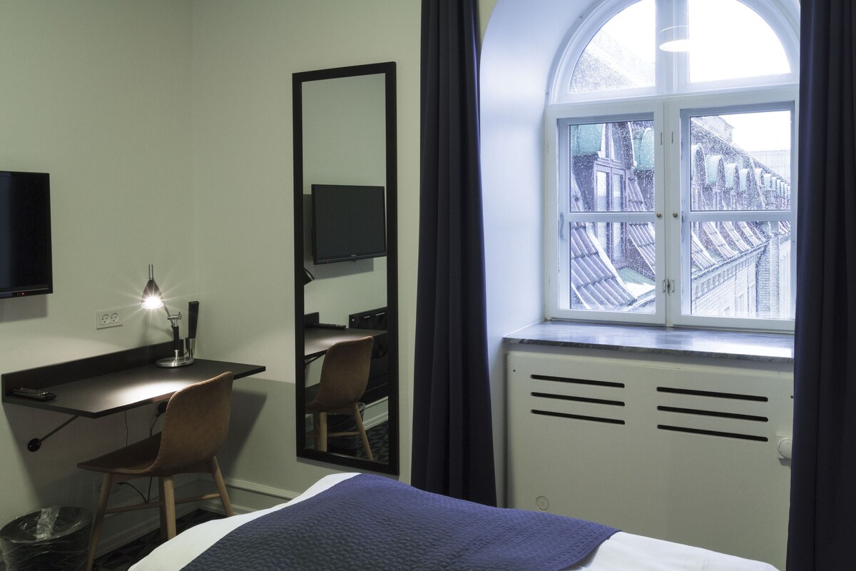 A hotel room with a bed, a desk with a chair, a TV, a window with curtains, and a radiator.