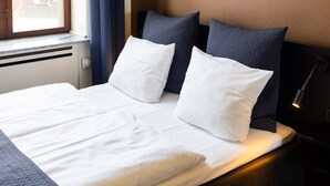 A neatly made bed with white linens and two dark pillows, a bedside lamp, and a window with curtains. - Milling Hotel Ritz (Aarhus)