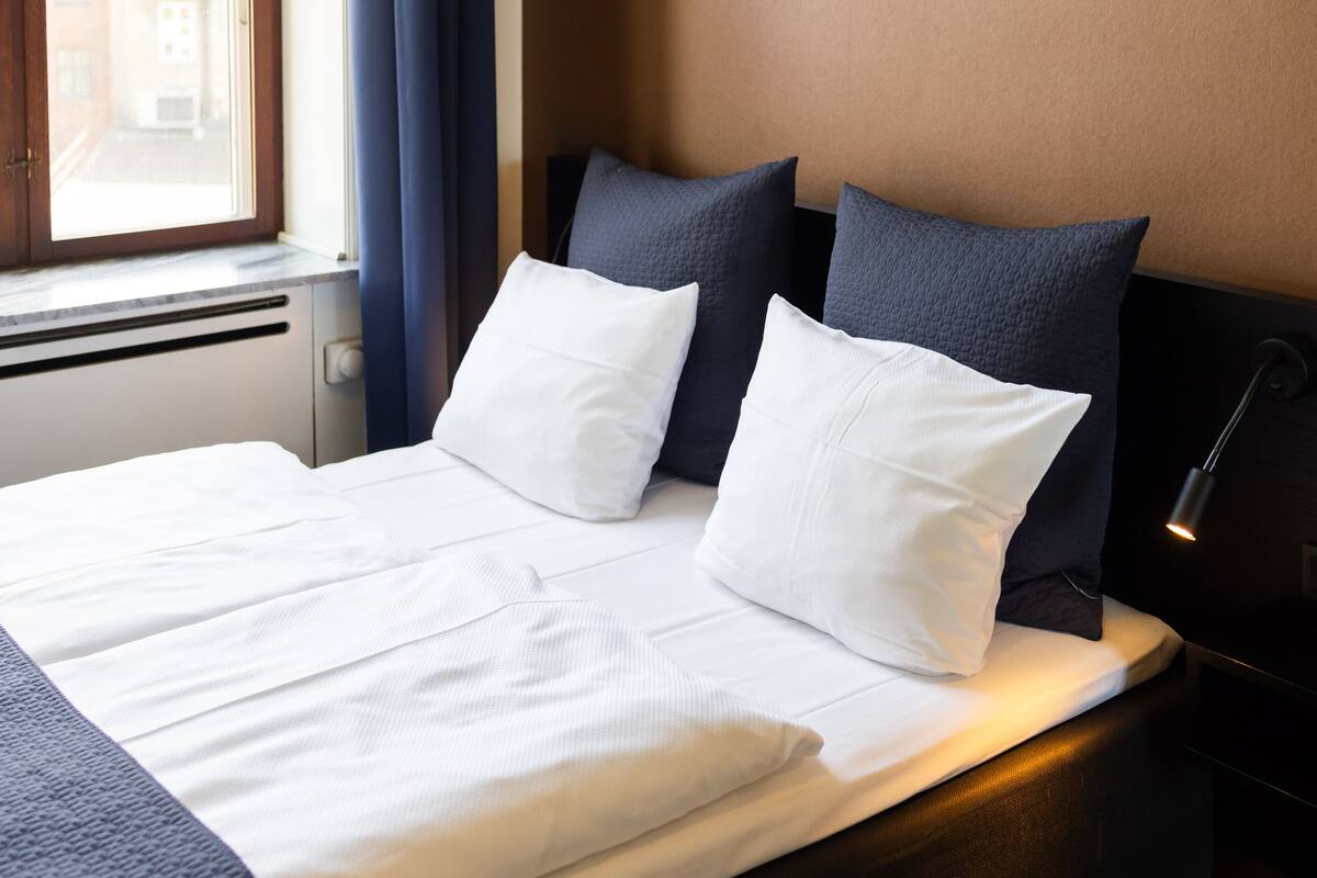 Een netjes opgemaakt bed met witte lakens en twee donkere kussens, een bedlampje en een raam met gordijnen.