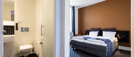 Una habitación de hotel con baño, una cama con colcha azul estampada y una pared de acento marrón.