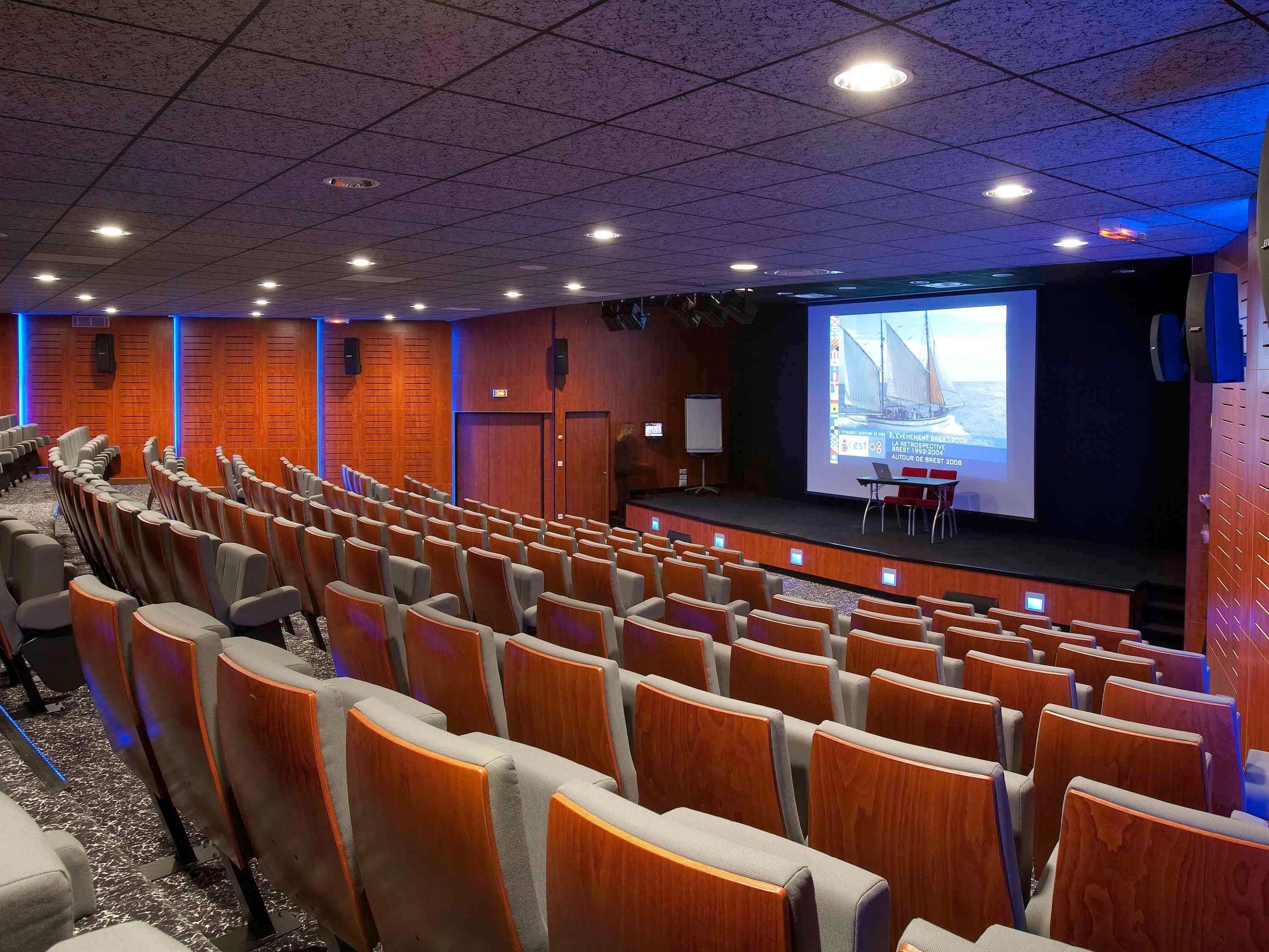 A conference room with rows of chairs, a stage with a presentation screen, wooden paneling, and ceiling lights.